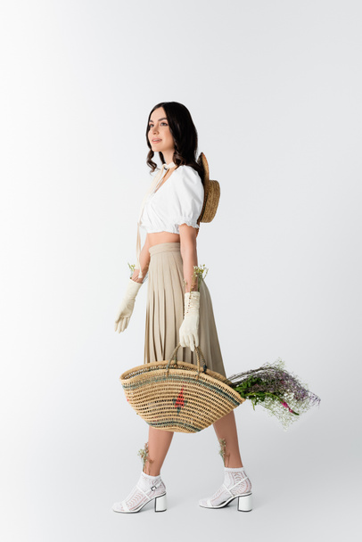 full length of happy woman in spring outfit holding straw bag with flowers on white - Фото, изображение