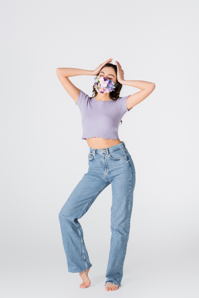 полная длина молодой женщины в кукурузном топе, джинсах и медицинской маске с цветущими цветами, позирующими на белом - Фото, изображение