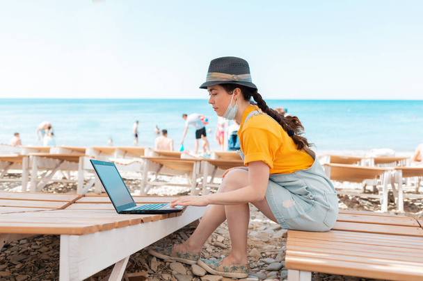 Szabadúszó. Egy nő orvosi maszkkal az arcán, ül egy széken, és gépel egy laptopon. A háttérben a tengerpart és a tenger. A távmunka fogalma világjárvány idején. - Fotó, kép