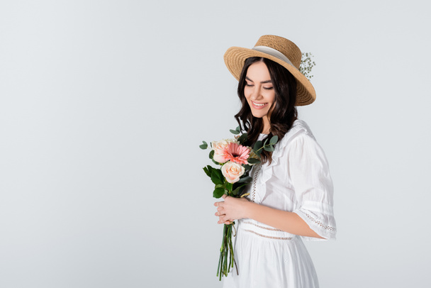 cheerful young woman in straw hat and dress holding bouquet of spring flowers isolated on white - Fotografie, Obrázek