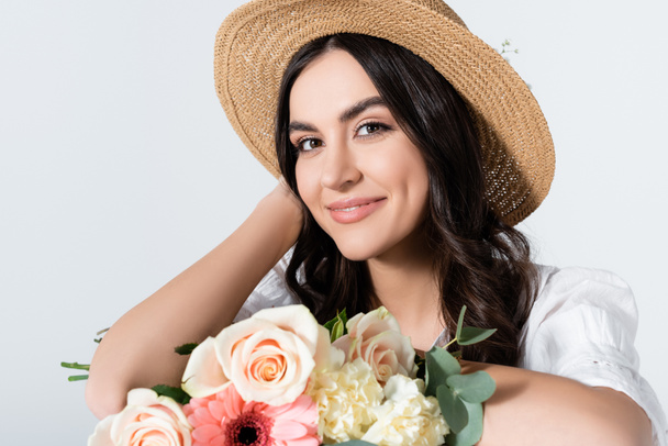 улыбающаяся молодая женщина в соломенной шляпе, держащая букет весенних цветов, изолированных на белом - Фото, изображение