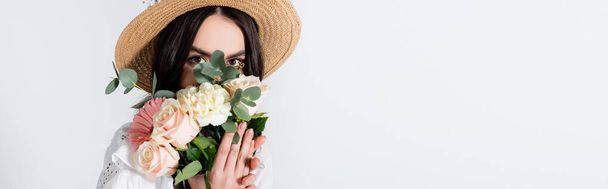 mladá žena v slamáku zakrývající tvář s kyticí jarních květin izolovaných na bílém, prapor - Fotografie, Obrázek
