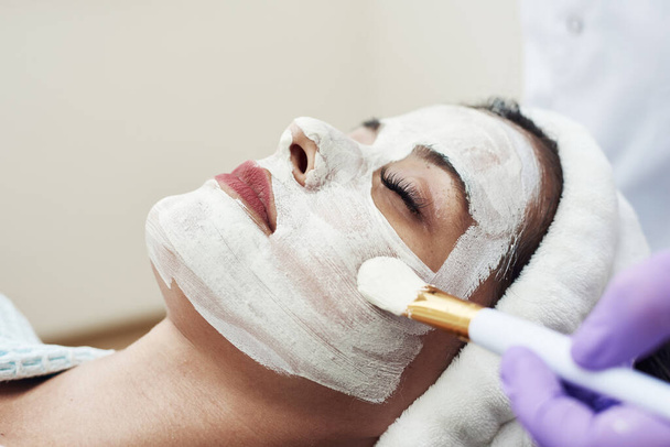 Una excelente esteticista aplica una máscara hidratante blanca anti-envejecimiento en la cara con un cepillo - Foto, Imagen