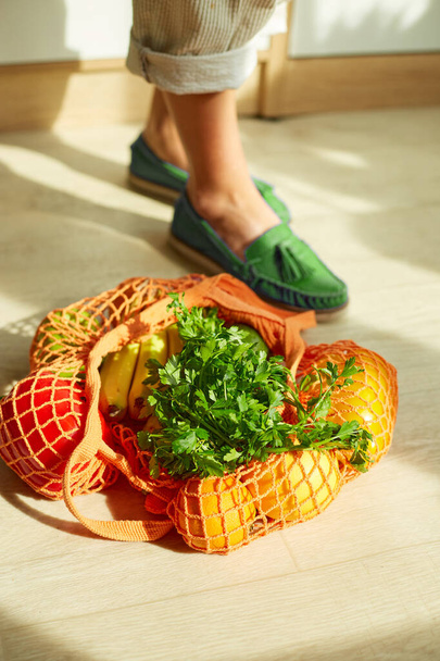 Shopping string grocery reusable mesh bag full of fresh fruits and vegetables on the floor at home, sunlight with top view of woman leg in green shoes. Concept healthy vegan eating, Zero waste. Space for text, Reusable eco friendly object - Photo, Image