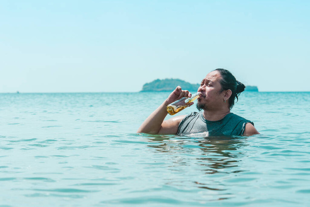 Ένας Κινέζος με μπλουζάκι πίνει ένα μπουκάλι μπύρα ενώ στέκεται στο στήθος βαθιά μέσα στο νερό σε μια παραλία στη μέση της ημέρας.. - Φωτογραφία, εικόνα