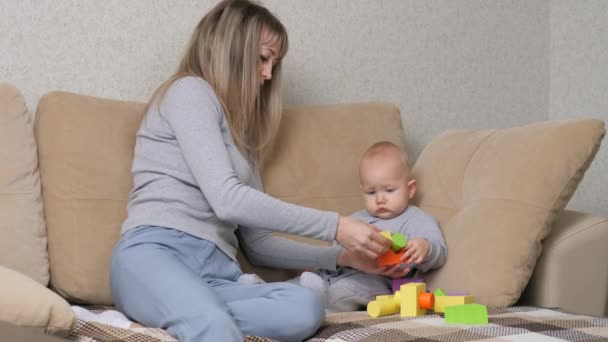 Bebek ve anne birlikte kanepede renkli küplerle oynuyorlar. Mutlu bir aile. Anne çocuklarının motor becerilerini geliştirir. Bir çocuğa oyun oynayarak öğretmek. Çocuklar için eğitici oyunlar - Video, Çekim