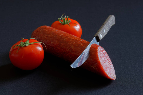 トマト、サラミ、暗めの背景に鋭いナイフ。ユダヤ教における男性割礼の概念と比喩。危険と痛み。閉鎖 - 写真・画像