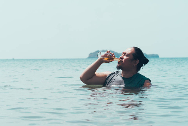 Ein fetter asiatischer Kerl trinkt eine Flasche Bier, während er mitten am Tag an einem Strand tief im Wasser steht. - Foto, Bild