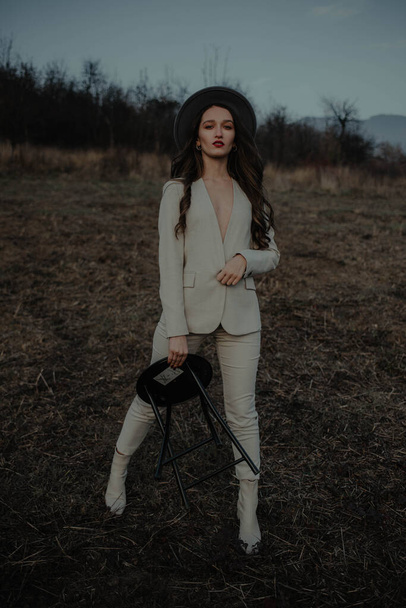 A full-body shot of a sexy confident woman with a trendy outfit standing in the dark forest holding a black minimalmetal chair - Photo, image