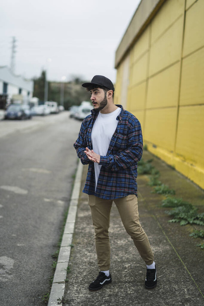 A stylish young European male wearing a snapback hat posing on background of a yellow building - Fotó, kép