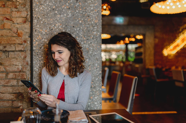 A portrait of a smiling young woman reading and typing on her phone while waiting for an order - Foto, afbeelding