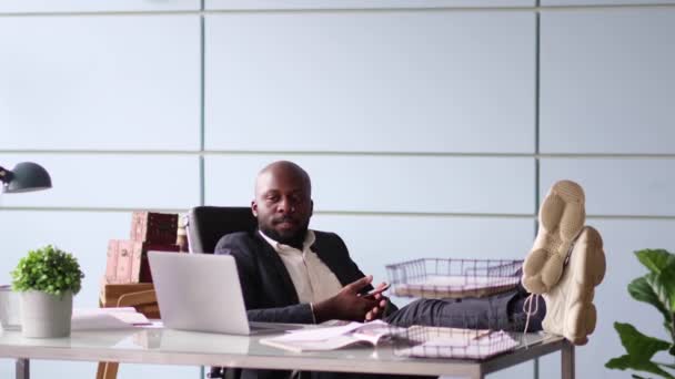 Nahaufnahme eines afroamerikanischen Geschäftsmannes, während die Füße auf dem Schreibtisch liegen. - Filmmaterial, Video