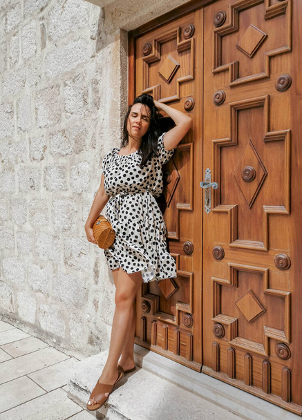 A confident Caucasian woman in a polka-dotted flowy dress posing against a decorative door - Photo, image