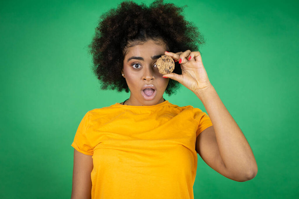 African american woman wearing orange casual shirt over green background holding healthy chocolate cookie over eye afraid and shocked with surprise and amazed expression, fear and excited face. - Photo, Image