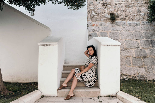 A confident Caucasian woman in a polka-dotted flowy dress sitting on white stairs outside - Foto, imagen