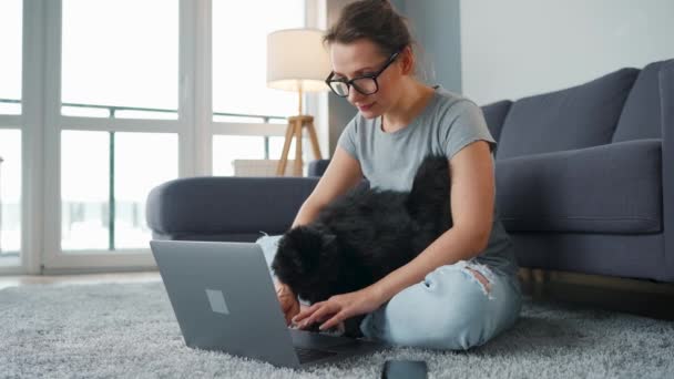 Femme vêtue de façon décontractée avec des lunettes s'assoit sur un tapis avec un ordinateur portable et caresse un chat noir moelleux tout en travaillant dans une chambre confortable. - Séquence, vidéo