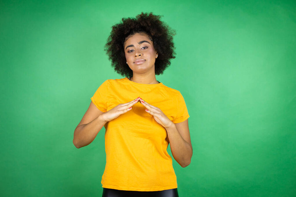 緑の背景にオレンジのカジュアルなシャツを着たアフリカ系アメリカ人女性が手を合わせて、指がリラックスして陽気に笑顔を交わした。成功と楽観的 - 写真・画像