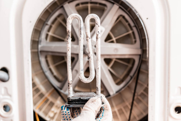 A worker holds a broken washing machine heating element covered in scale. Close up. In the background, the drum of the washing machine. Repair concept. - Photo, Image