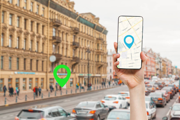 A női kéz egy okostelefont tart online térképalkalmazással. A háttérben egy elmosódott városi utcában a forgalom, egy zöld hely ikon.Fogalom az online navigáció és a GPS. - Fotó, kép