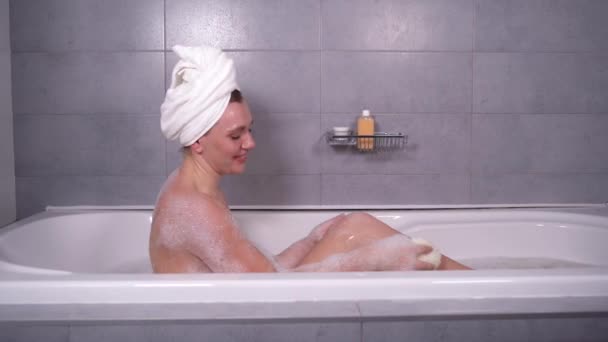 young caucasian woman is sitting in a bath with a towel on her head. A lot of penny. Rubs the body with a body sponge. Vacation home concept. - Footage, Video