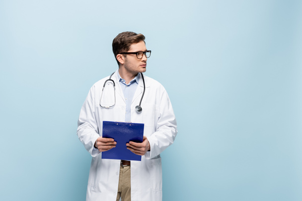 jeune médecin en lunettes et manteau blanc tenant presse-papiers et regardant loin sur bleu - Photo, image