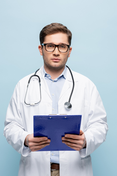 giovane medico in occhiali e mantello bianco che tiene appunti e guarda la fotocamera isolata sul blu - Foto, immagini