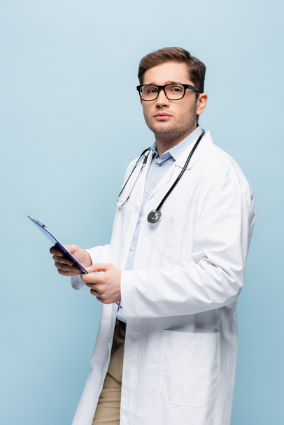 doctor in glasses and white coat holding clipboard isolated on blue - Photo, Image