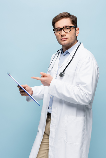 joven médico en gafas y abrigo blanco apuntando con el dedo al portapapeles aislado en azul - Foto, Imagen