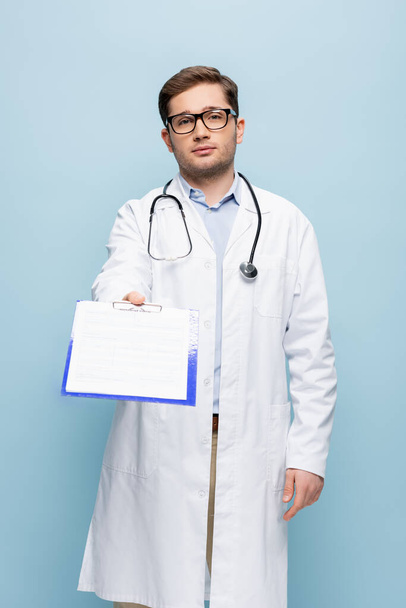doctor in glasses and white coat giving clipboard with medical card lettering isolated on blue - Photo, Image