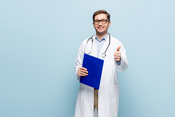 cheerful doctor in glasses and white coat pointing with finger at clipboard and showing thumb up on blue - Photo, Image
