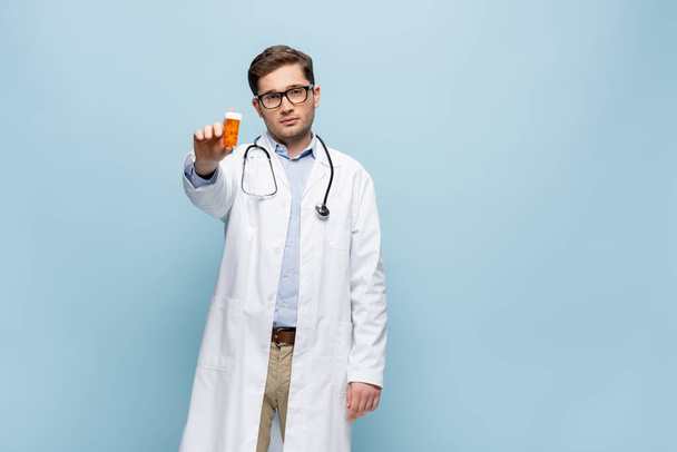 Arzt in Brille und weißem Mantel, Flasche mit Medikamenten isoliert auf blauem Grund - Foto, Bild