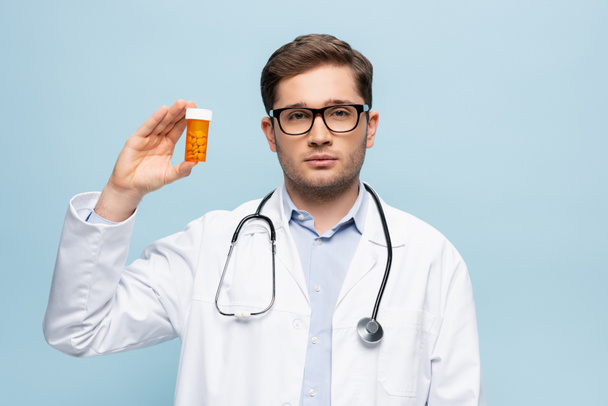 joven doctor en gafas y abrigo blanco sosteniendo botella con medicamentos aislados en azul - Foto, imagen