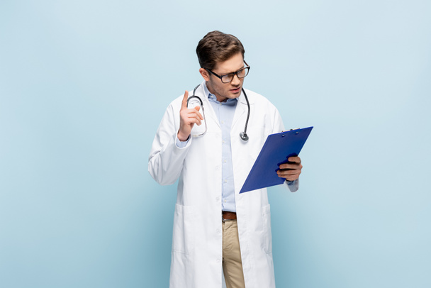 Arzt mit Brille und weißem Mantel zeigt Ideenschild und blickt auf Klemmbrett auf blauem Grund - Foto, Bild