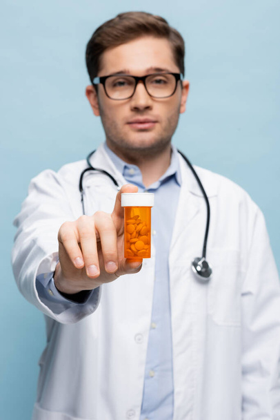 médecin flou dans des lunettes et manteau blanc tenant bouteille avec des médicaments isolés sur bleu - Photo, image