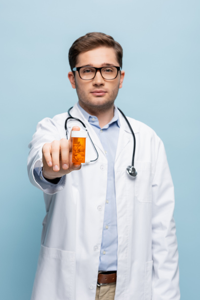 verschwommener junger Arzt mit Brille und weißem Mantel, Flasche mit Medikamenten isoliert auf blauem Grund - Foto, Bild
