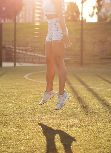 Fitness femme avec des jambes musclées porte un soutien-gorge de sport et des shorts sautant dans la cour de sport dans la journée ensoleillée - Photo, image