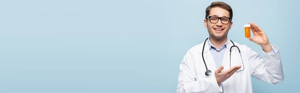 médecin heureux dans des lunettes et manteau blanc pointant avec bouteille à main avec des médicaments isolés sur bleu, bannière - Photo, image