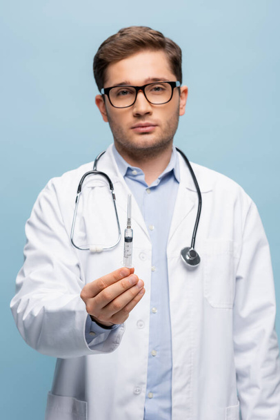 Arzt mit Brille und weißem Kittel hält Spritze in Impfstoff isoliert auf blau - Foto, Bild