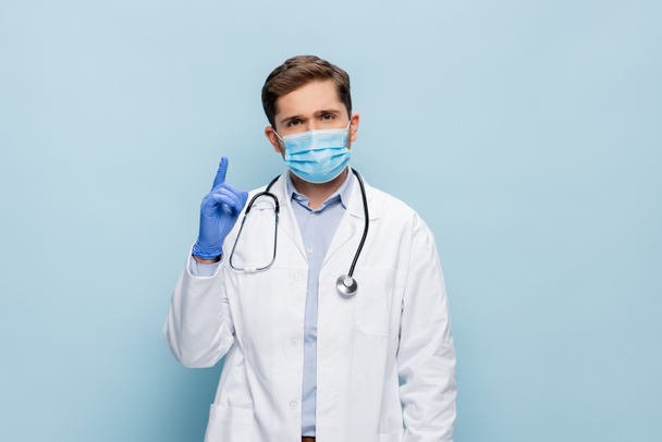 médico en máscara médica y bata blanca con estetoscopio apuntando hacia arriba con el dedo aislado en azul - Foto, Imagen