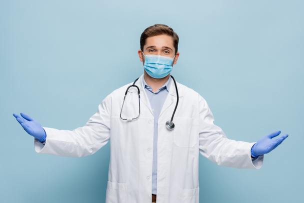 médico en máscara médica y bata blanca con estetoscopio de pie con las manos extendidas en azul - Foto, Imagen