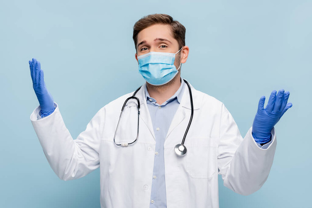 молодой врач в медицинской маске и белом халате, стоящий с протянутыми руками, изолированными на голубом - Фото, изображение