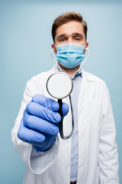 rozmazaný lékař v lékařské masce a bílý kabát drží stetoskop izolované na modré - Fotografie, Obrázek