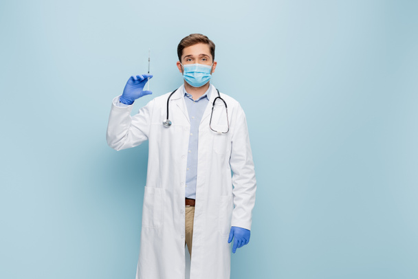 young doctor in medical mask and white coat with stethoscope holding syringe on blue - Photo, Image