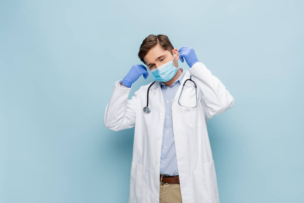joven médico de bata blanca con estetoscopio con máscara médica en azul - Foto, imagen