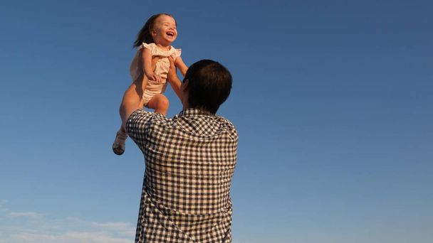 Dítě si hraje a chce létat jako letadlo. Táta hodí svou šťastnou dceru na modrou oblohu. Rodinné cestování. Otec a malé dítě si hrají, smějí se, objímají se. Rodinná procházka v parku v létě, víkend - Fotografie, Obrázek