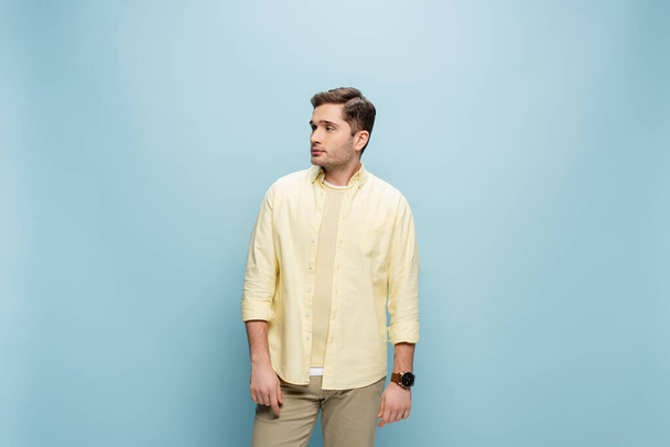 νεαρός άνδρας με κίτρινο πουκάμισο κοιτάζει αλλού και στέκεται απομονωμένος στο μπλε - Φωτογραφία, εικόνα