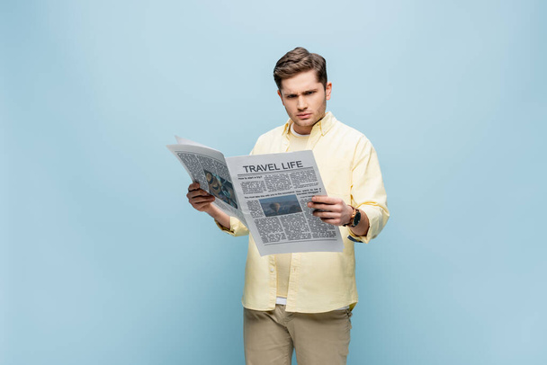 Tişörtlü, seyahat gazetesi okuyan ciddi bir genç adam mavi üzerine izole edilmiş. - Fotoğraf, Görsel