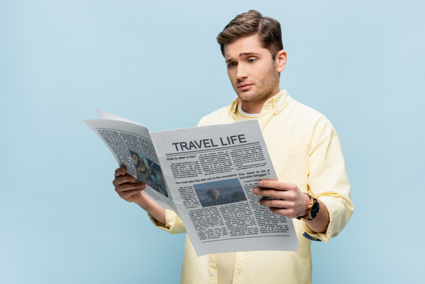 sceptische jongeman in shirt lezen reiskrant geïsoleerd op blauw - Foto, afbeelding