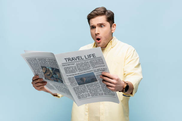 Şok olmuş genç adam mavi üzerine izole edilmiş seyahat gazetesi okuyor. - Fotoğraf, Görsel