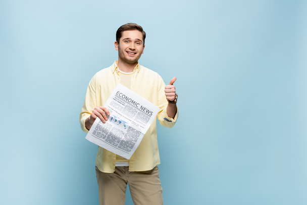 uśmiechnięty mężczyzna w koszuli trzyma gazetę i pokazuje kciuk w górze odizolowany na niebiesko - Zdjęcie, obraz